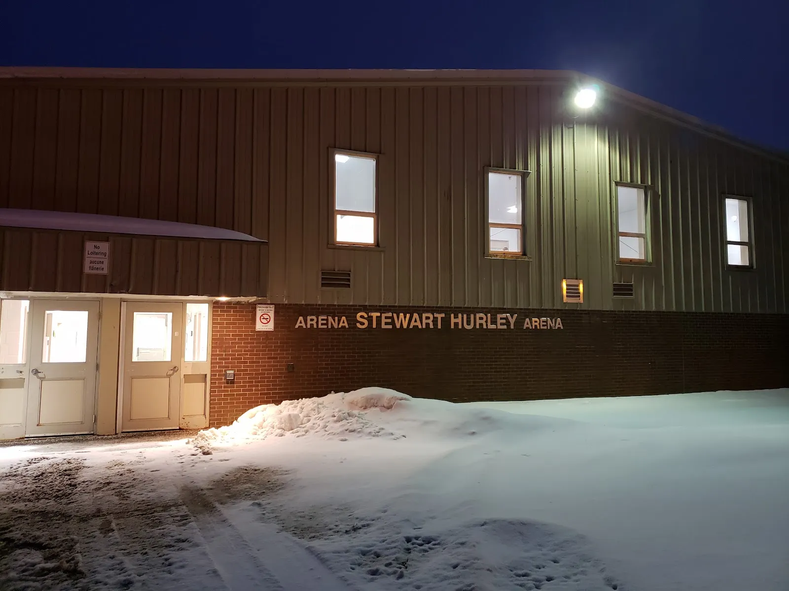 The brick entrance wall and metal-clad side of Stewart Hurley Arena in Saint John at dusk, with the arena name in lettering beside the doors — Stewart Hurley Arena