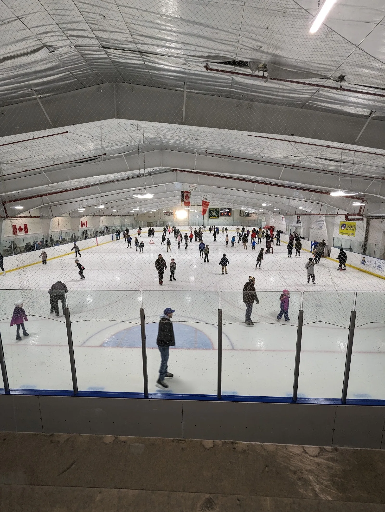 A crowded public session far down a wide sheet, many skaters spread along the boards beneath a ribbed roof with a banner at the far end. — Stewart Hurley Arena