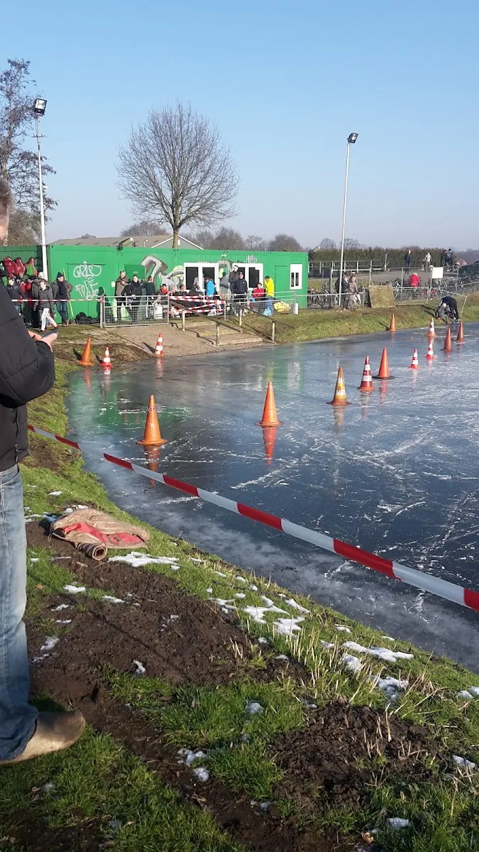 The green clubhouse beside the natural ice of IJsbaan de Grote Maot in Bathmen, with traffic cones and tape along the ice edge – IJsbaan De Grote Maot Bathmen