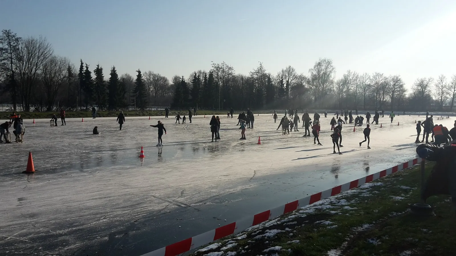 Skaters at a distance on the wide natural ice of IJsbaan de Grote Maot in Bathmen, with the red and white tape along the edge and trees beyond – IJsbaan De Grote Maot Bathmen