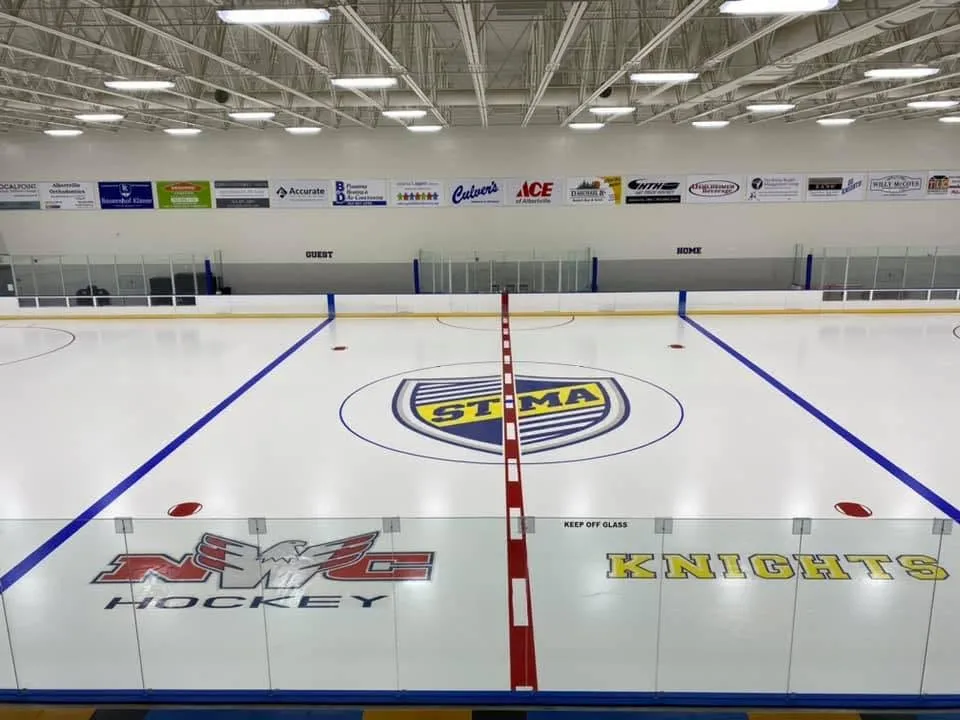 Full ice sheet looking down the center line toward the far end, with the STMA logo at center ice and "KNIGHTS" lettering on the boards. — STMA Ice Arena (St. Michael-Albertville Arena)