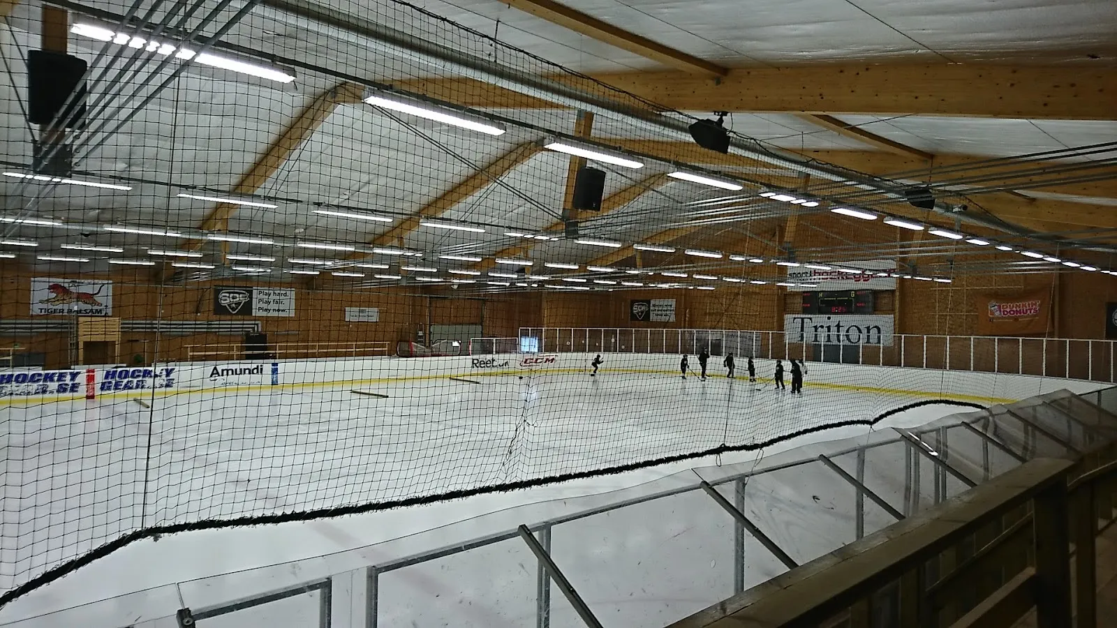 Isen i Stockhagens ishall sedd från läktaren genom skyddsnätet, med några skridskoåkare långt borta vid sargen under ett tak av trä – Stockhagens ishall