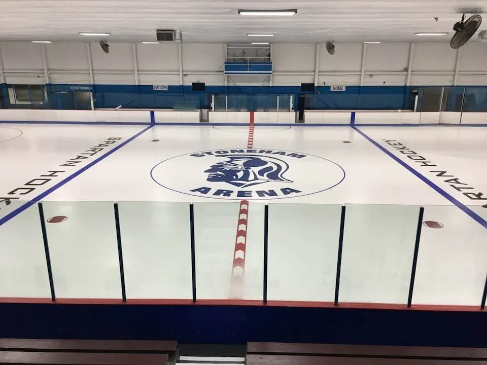 Full NHL-size ice sheet at Stoneham Arena, empty, with a Stoneham Arena crest painted at center ice and HOME/VISITORS bench doors along the boards. — Stoneham Arena
