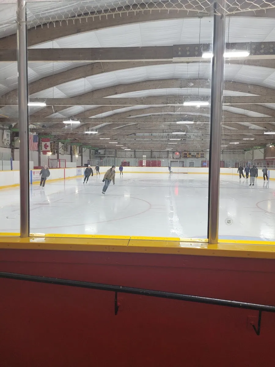 The ice during a public skate, seen through the glass from the red-walled stands under the arched wooden roof beams. — Stoney Creek Arena