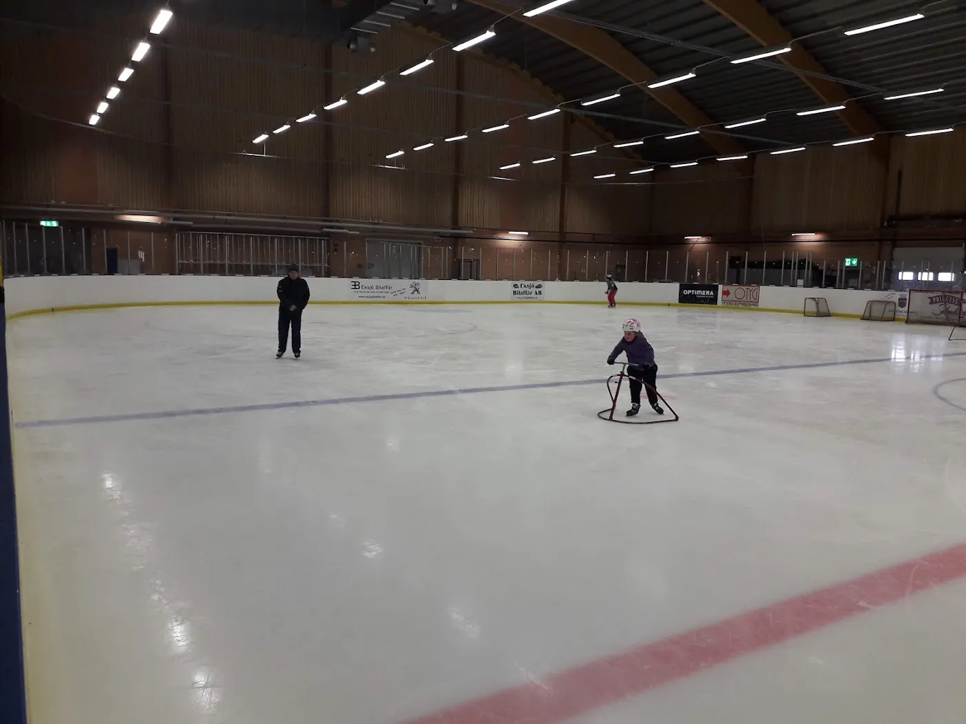 Isen i Storegårdshallen i Eksjö, en bred hall med trätak där isen är nästan tom och ett barn med åkstöd och en vuxen syns långt bort vid sargen – Storegårdshallen