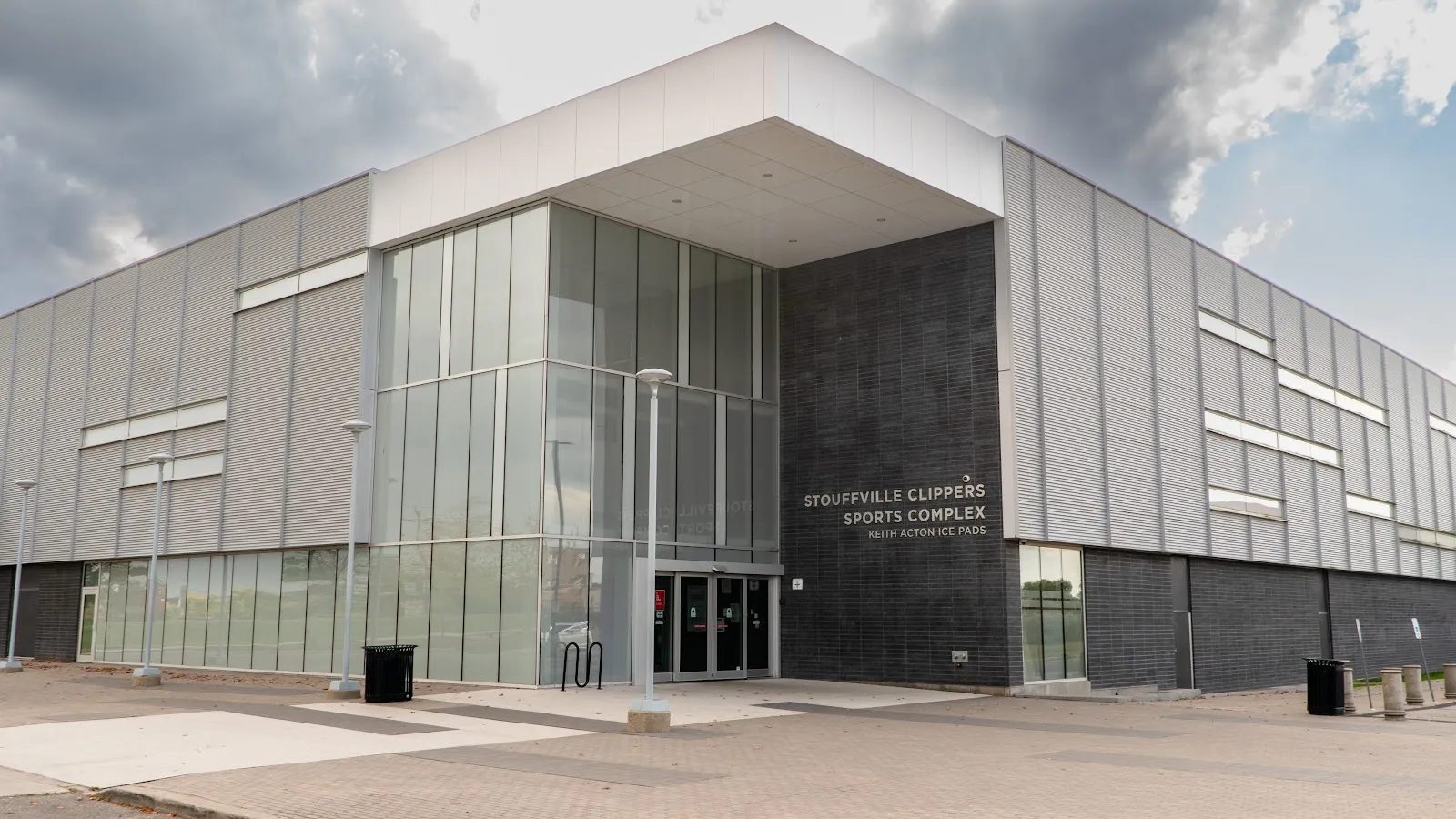 La torre de entrada de vidrio del complejo junto a un muro oscuro con la leyenda Stouffville Clippers Sports Complex, con muros de paneles gris claro a ambos lados. — Stouffville Clippers Sports Complex