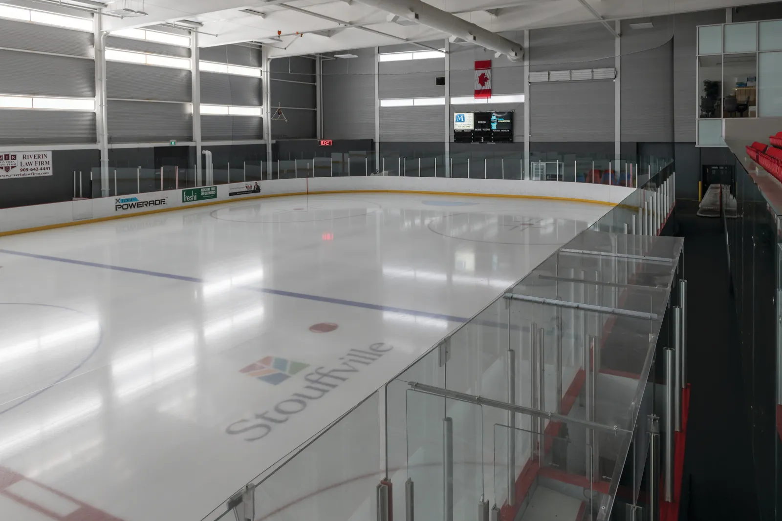 El hielo vacío con el logotipo de Stouffville en el centro, visto desde arriba del vidrio junto a los asientos rojos, con un marcador y una bandera en el muro del fondo. — Stouffville Clippers Sports Complex
