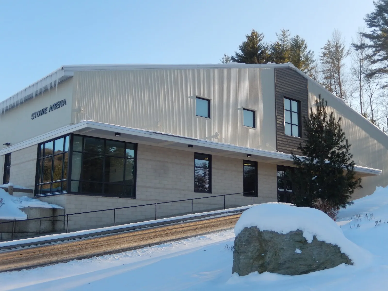 La arena de Park Street en invierno, con su nombre en letras oscuras sobre la nave metálica clara de la pista, encima de un vestíbulo vidriado y una roca nevada — Stowe Arena