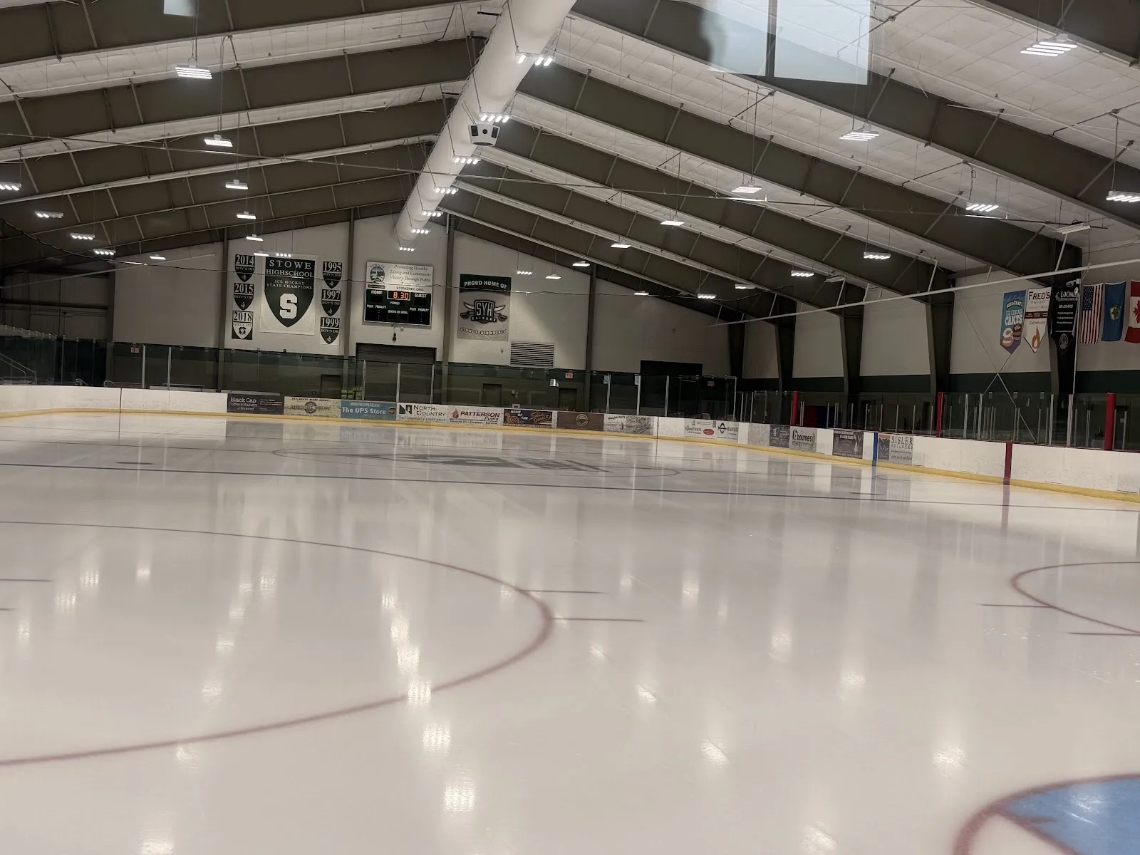 Una pista vacía con tenues círculos bajo un techo de madera y acero del que cuelgan banderines y estandartes, con vallas y bancas blancas en la pared del fondo. — Stowe Arena
