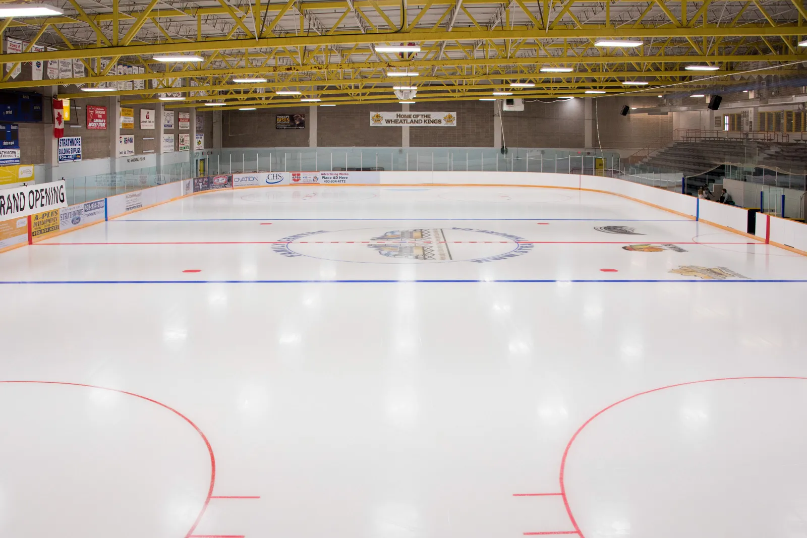 La pista Gold vacía (de tamaño olímpico) bajo armaduras amarillas de acero, con una manta de Grand Opening y anuncios de patrocinadores en el lado opuesto. — Strathmore Family Centre Arena