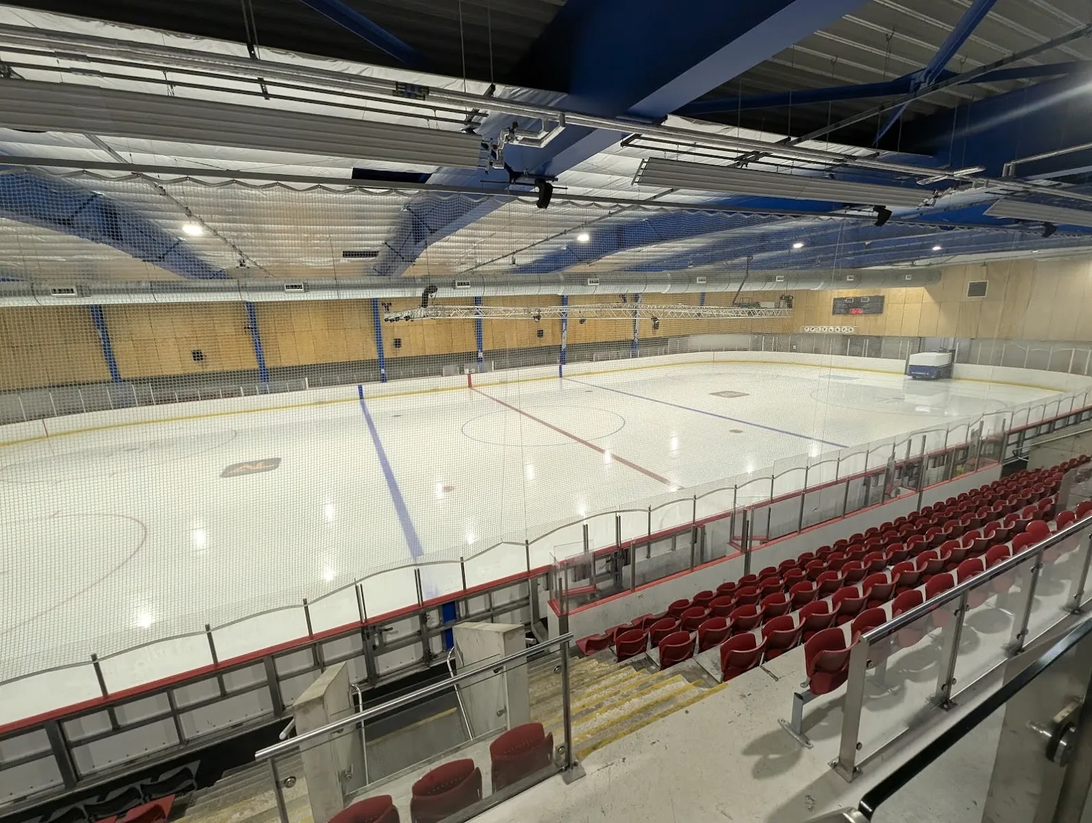The empty ice pad seen from the upper seating, with red tip-up seats along one side and a timber-lined roof above. — Streatham Ice and Leisure Centre