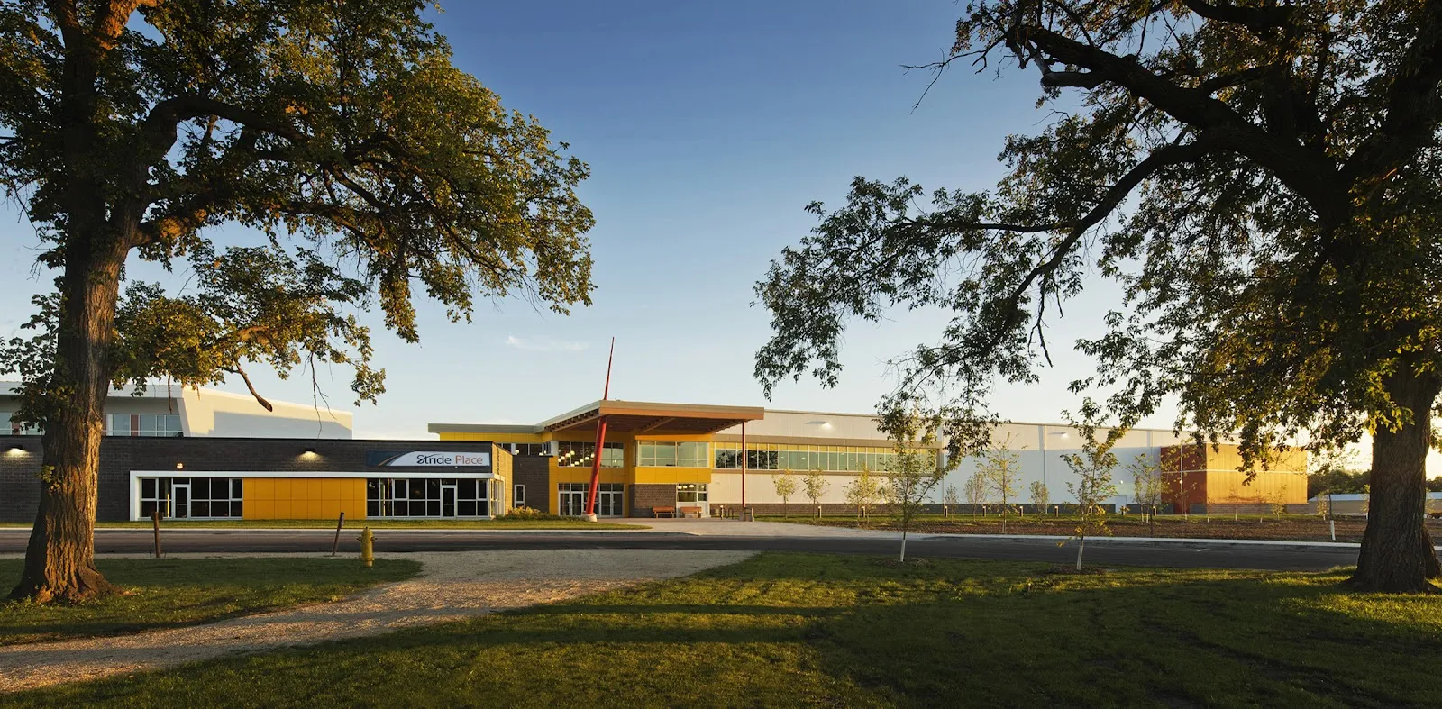 The long low front of Stride Place with its yellow and dark panels and glass entrance, seen across a lawn between two large trees at sunset. — Stride Place