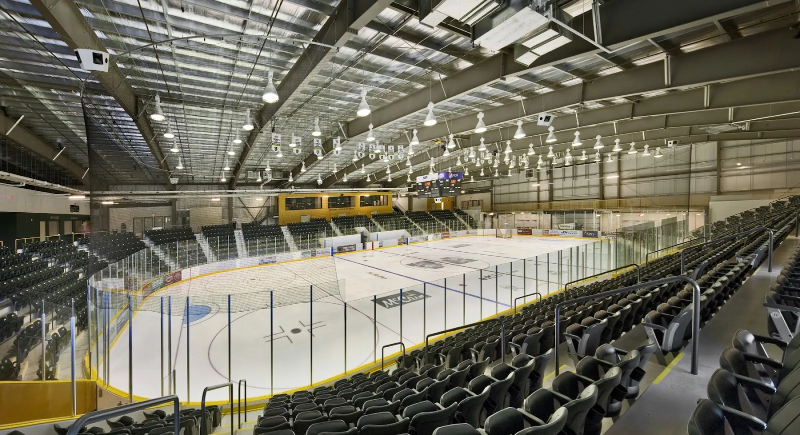 The main arena bowl from the upper seats: rows of dark seating on both sides, glass around the empty ice, and exposed roof trusses with lights. — Stride Place