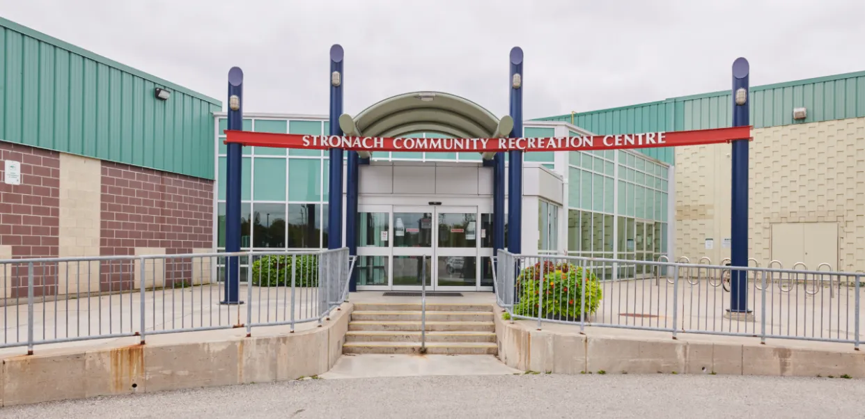 The glass-fronted entrance of the Stronach Community Recreation Centre in London, with a red banner above the doors and steps up from the sidewalk. — Stronach Arena
