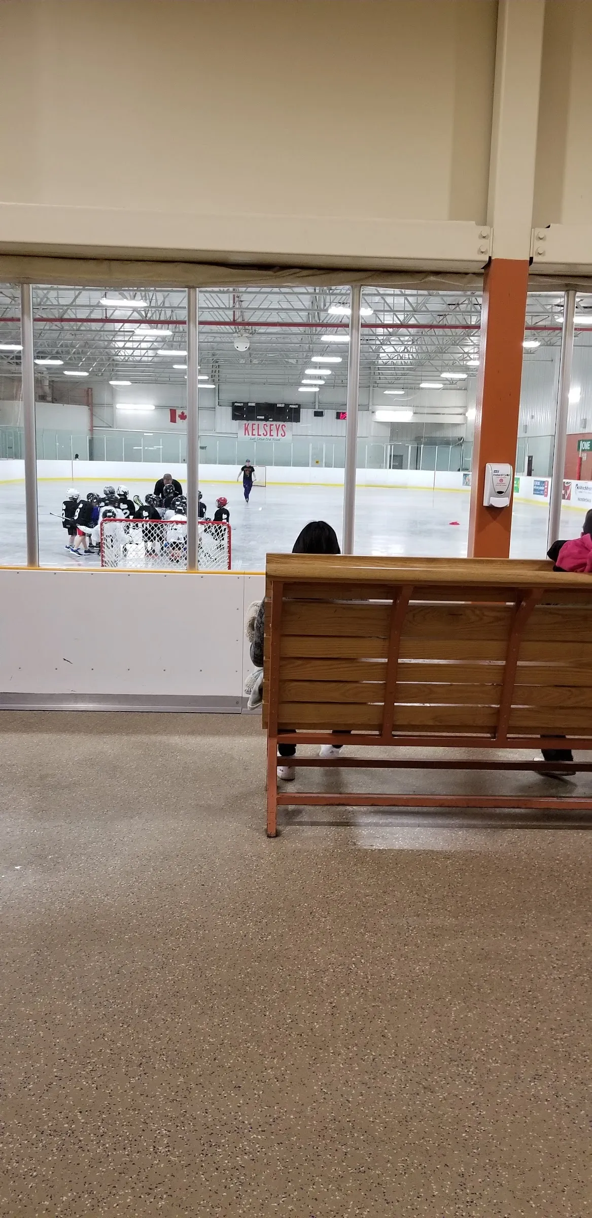 El hielo visto a través de las ventanas del área de observación, con una práctica de hockey juvenil en la portería del fondo, una bandera de Canadá y una manta de Kelseys sobre las vallas. — Stronach Arena