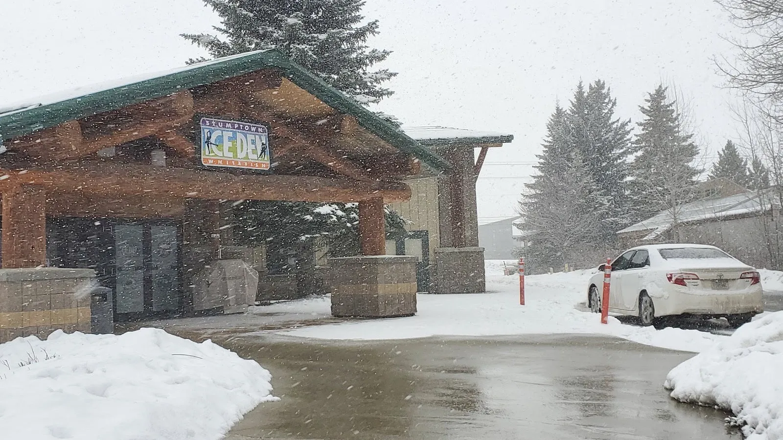 La entrada de Stumptown Ice Den mientras cae la nieve, con su letrero bajo un techo de pórtico de troncos con pilares de piedra — Stumptown Ice Den