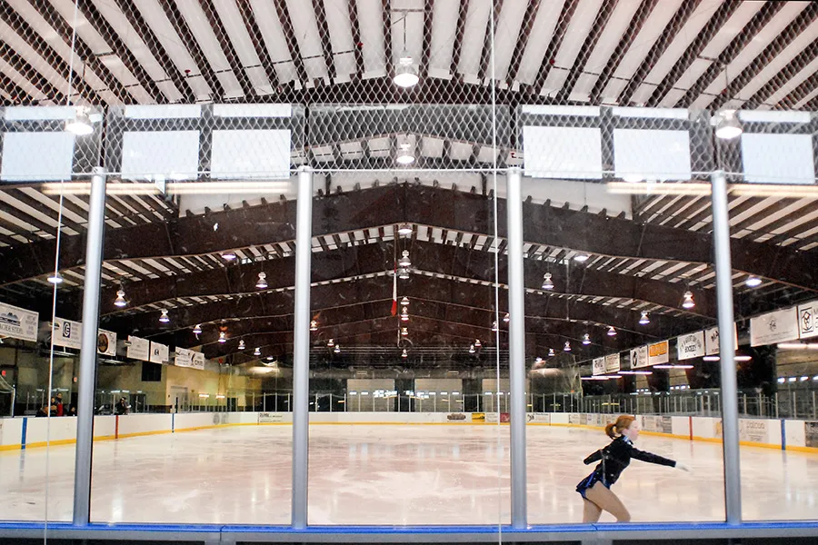 Pista de hielo vacía bajo un techo en arco con armaduras a la vista, vista a través del cristal junto a la pista, con un solo patinador practicando patinaje artístico cerca de las vallas y mantas de patrocinadores en la pared del fondo. — Stumptown Ice Den
