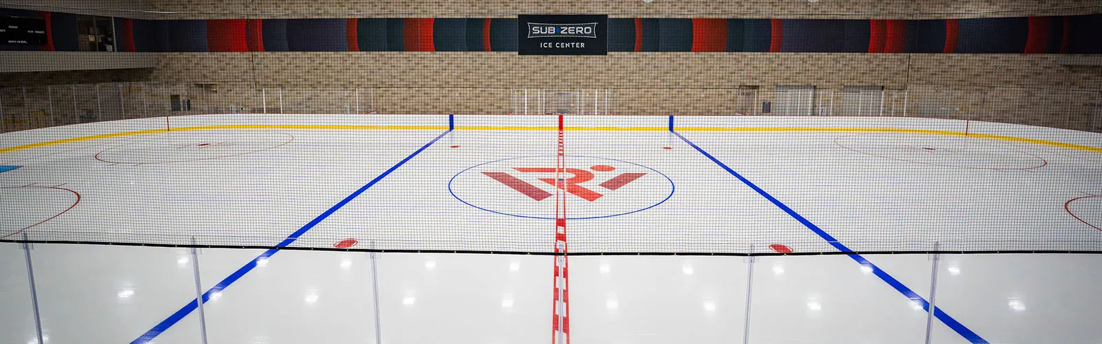 Pista de hielo vacía vista de un extremo a otro desde las vallas, con un letrero de "SUB-ZERO ICE CENTER" en la pared sobre la pista y un logotipo rojo de UW pintado en el centro de la pista. — Sub-Zero Ice Center