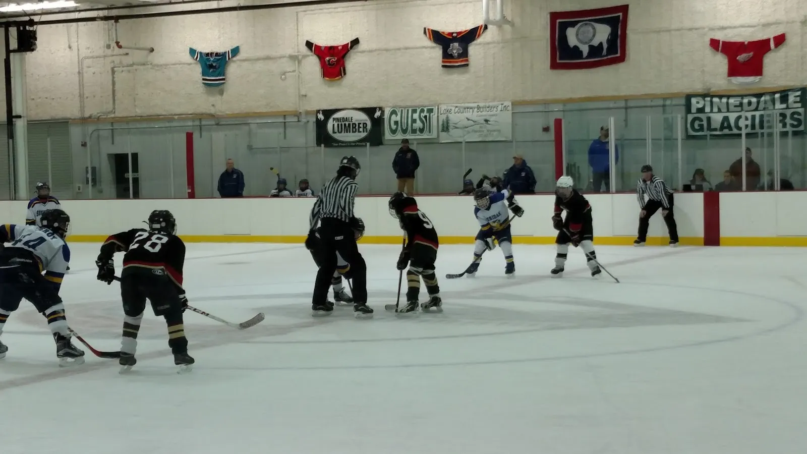 Youth hockey game at a faceoff, with Pinedale Glaciers boards and team jerseys hanging along the far wall of the arena. — Sublette County Ice Arena