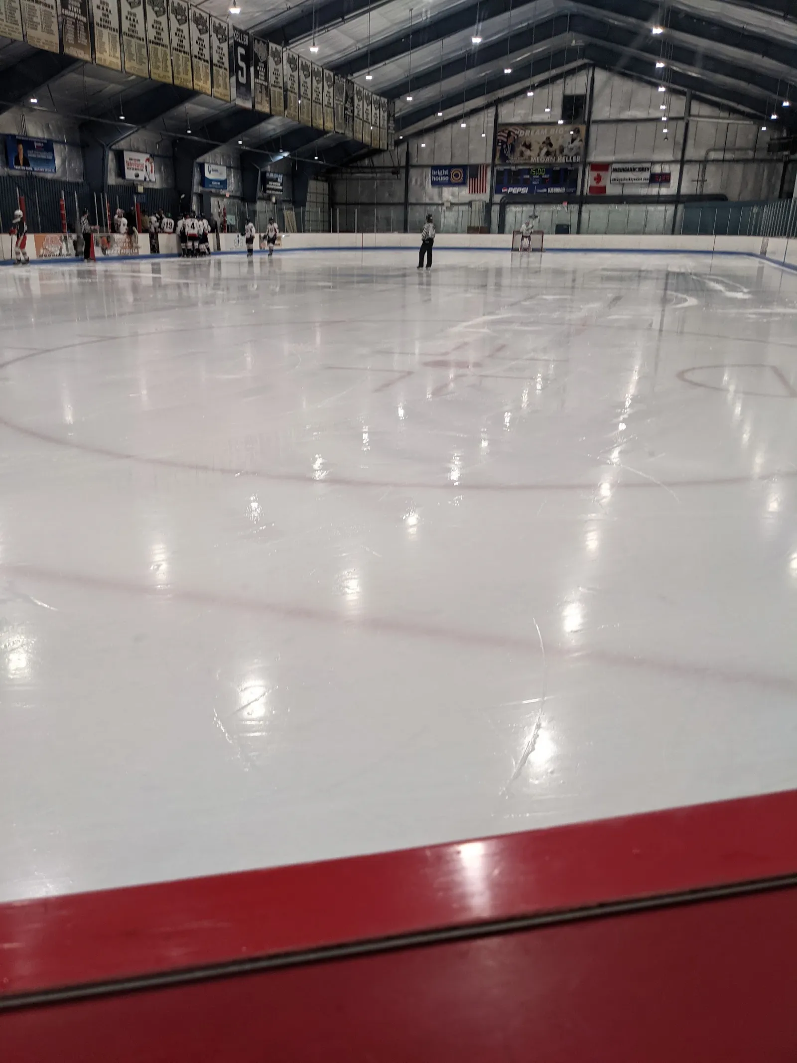 The ice seen from the red boards, with a youth team gathered at the far end, banners on the walls and a dark steel-truss roof. — Suburban Ice Farmington Hills