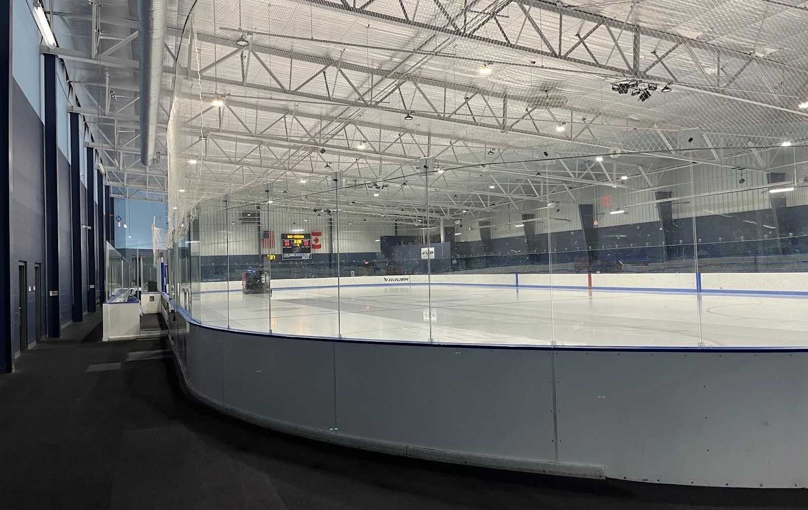 A Suburban Ice Macomb sheet seen past the curved glass from the concourse, freshly resurfaced, with the scoreboard and flags at the far end — Suburban Ice Macomb