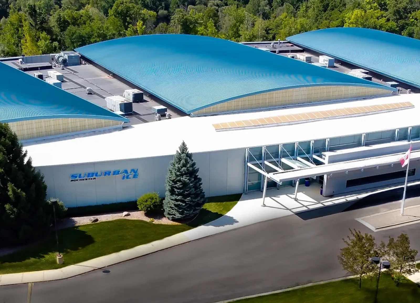 Aerial view of the arena's curved blue roof and white facade with a large 'Suburban Ice Rochester' wall sign over the entrance canopy. — Suburban Ice Rochester