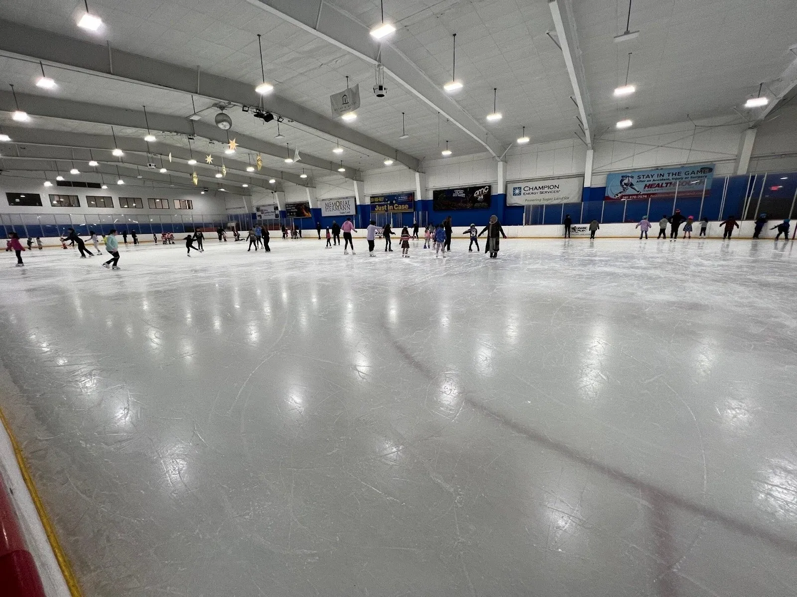 A wide sheet of grey-white ice with players spread along the far boards, a long steel-trussed roof and sponsor boards behind them. — Sugar Land Ice and Sports Center