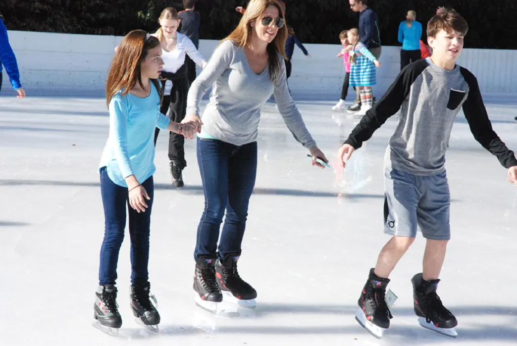 A parent helping a toddler with a skating aid across the sunlit outdoor ice at Sugar Mountain Resort, with other skaters behind. — Sugar Mountain Ice Skating Rink