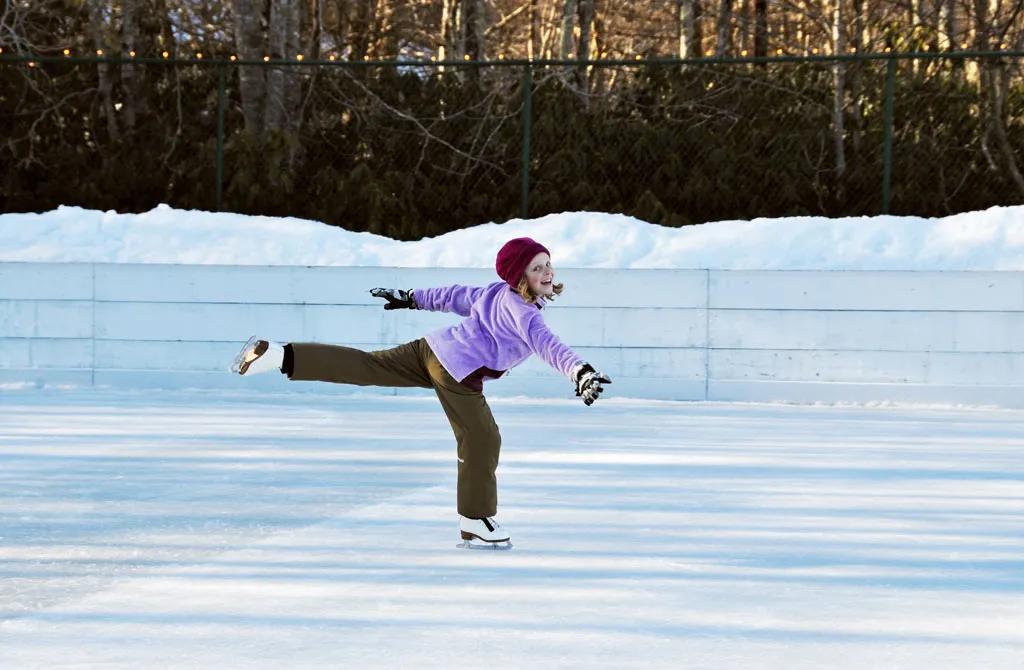 A skater gliding in an arabesque across the clear refrigerated ice, with snow banks and bare winter trees beyond the rink boards. — Sugar Mountain Ice Skating Rink