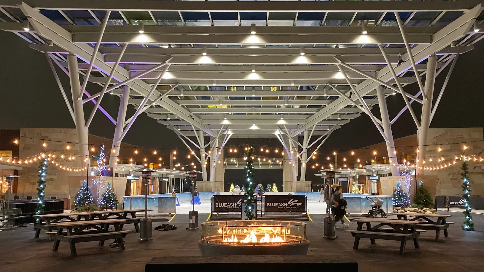 The glass-roofed canopy at night with a fire pit and picnic tables in front, the lit rink and holiday trees beneath it. — Ice Rink at Summit Park