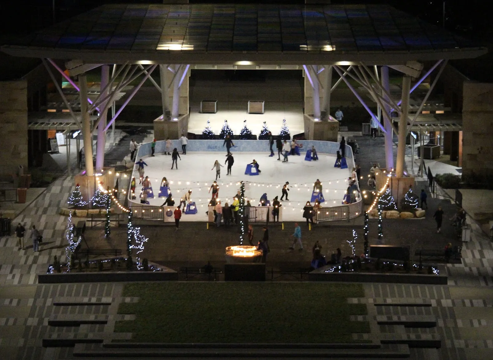 The whole rink seen from above under the canopy's steel columns, skaters circling the ice with a lit stage behind. — Ice Rink at Summit Park