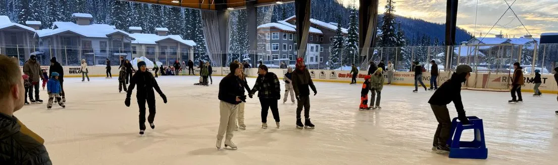 Skaters on the ice of the covered open-air Sun Peaks arena, with the village buildings and mountain slopes behind. — Sun Peaks Arena (Sun Peaks Centre)