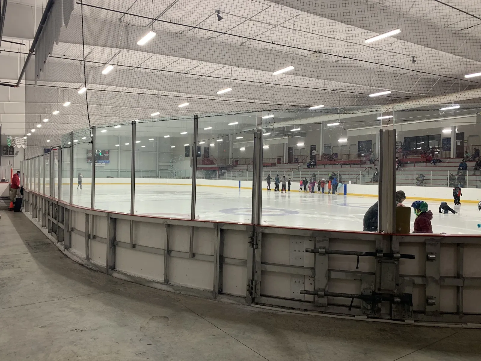 Una amplia vista de la pista desde detrás de las vallas durante una sesión de patinaje, con patinadores por todo el hielo y las gradas a lo largo del lado opuesto. — Sun Prairie Ice Arena