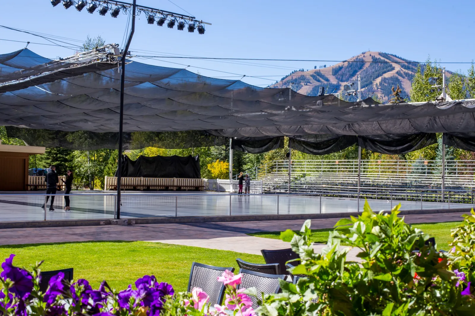 The outdoor rink under its shade canopy on a summer day, with a few distant skaters on the ice, Bald Mountain behind and flowers in the foreground. — Sun Valley Resort Ice Rink