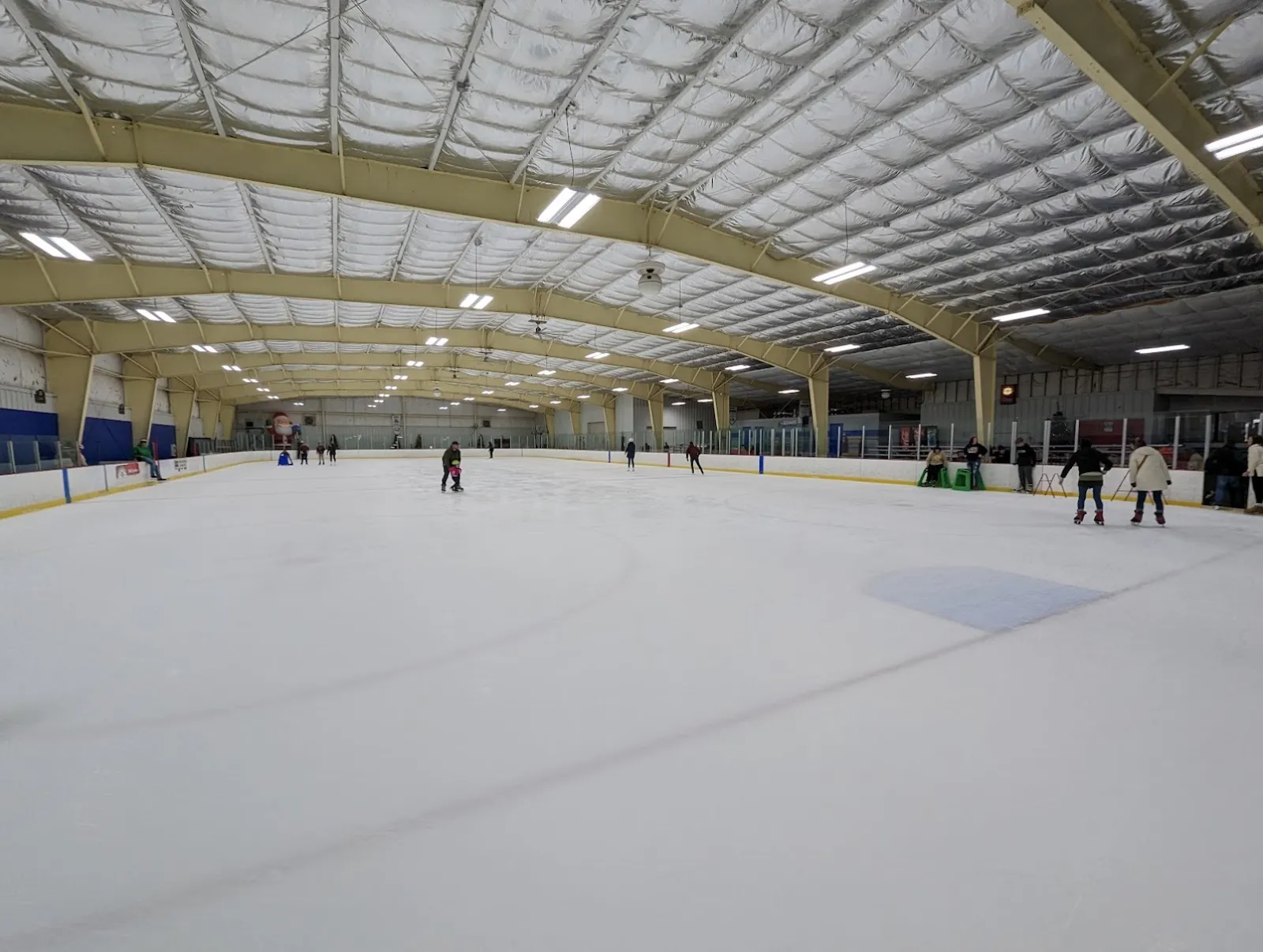 La pista techada vista desde las bandas, con unos pocos patinadores a lo lejos bajo un techo de arcos de acero con cielo raso aislado. — Sunbury Ice Rink
