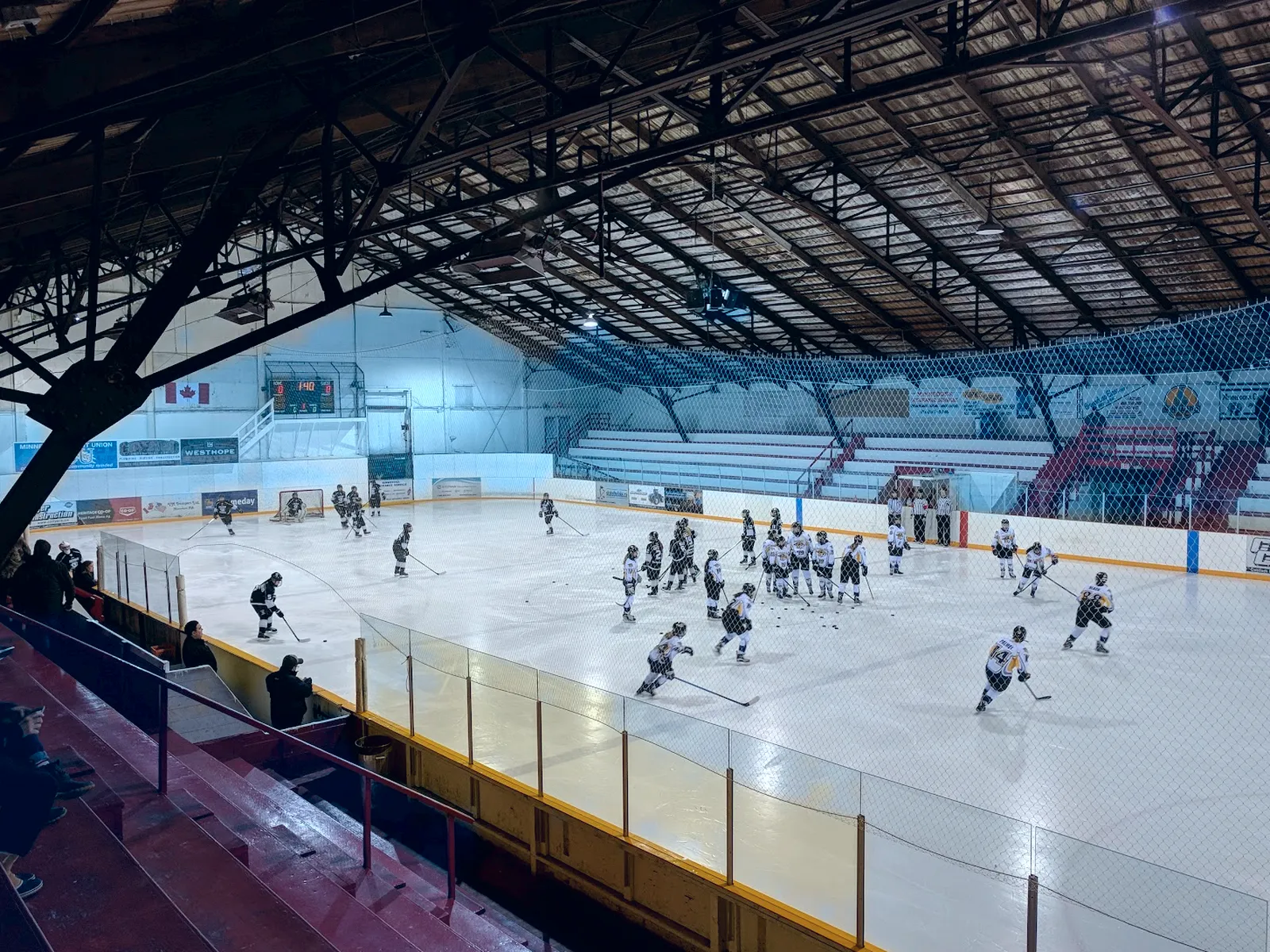 The ice from the red wooden bleachers under the arena's dark timber roof, with teams warming up and banners on the far wall. — Sunrise Credit Union Centre (Minnedosa)
