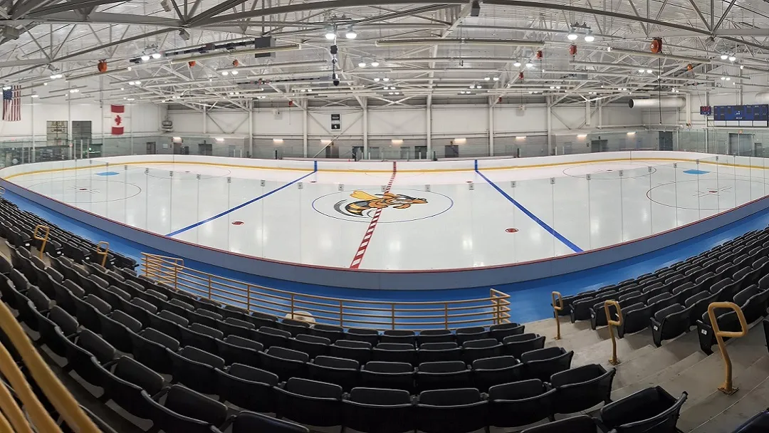 La pista vacía vista desde lo alto de las gradas, con el logo del avispón en el centro del hielo, filas de asientos oscuros en el lado cercano y las armaduras de acero del techo y las luces arriba. — SUNY Broome Ice Center