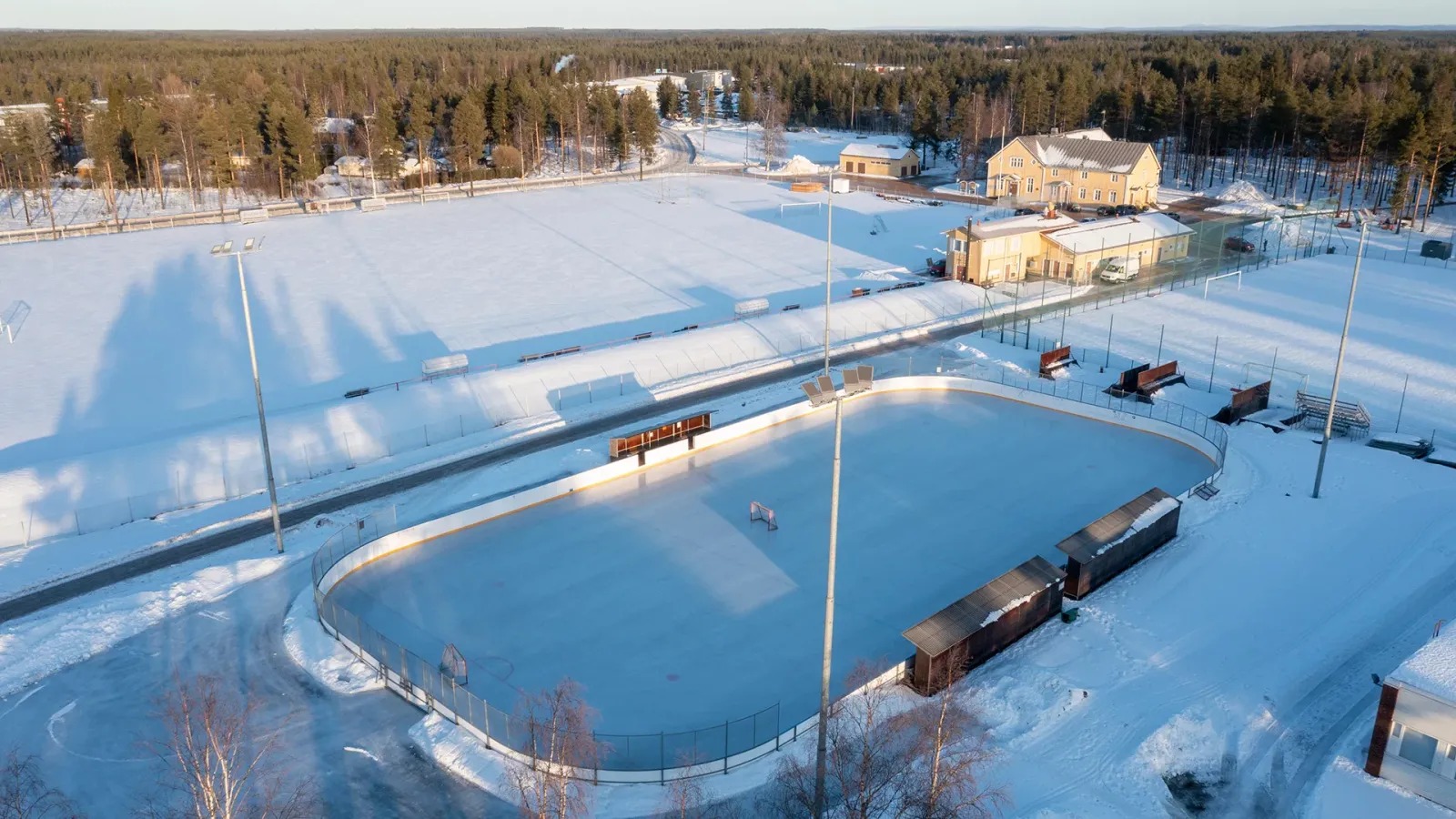 Aerial view of the Suojalinna rink in Pudasjärvi in snow: a boarded ice rink with floodlight poles and low buildings beside a snow-covered sports field. — Suojalinnan kaukalo (Pudasjärvi)