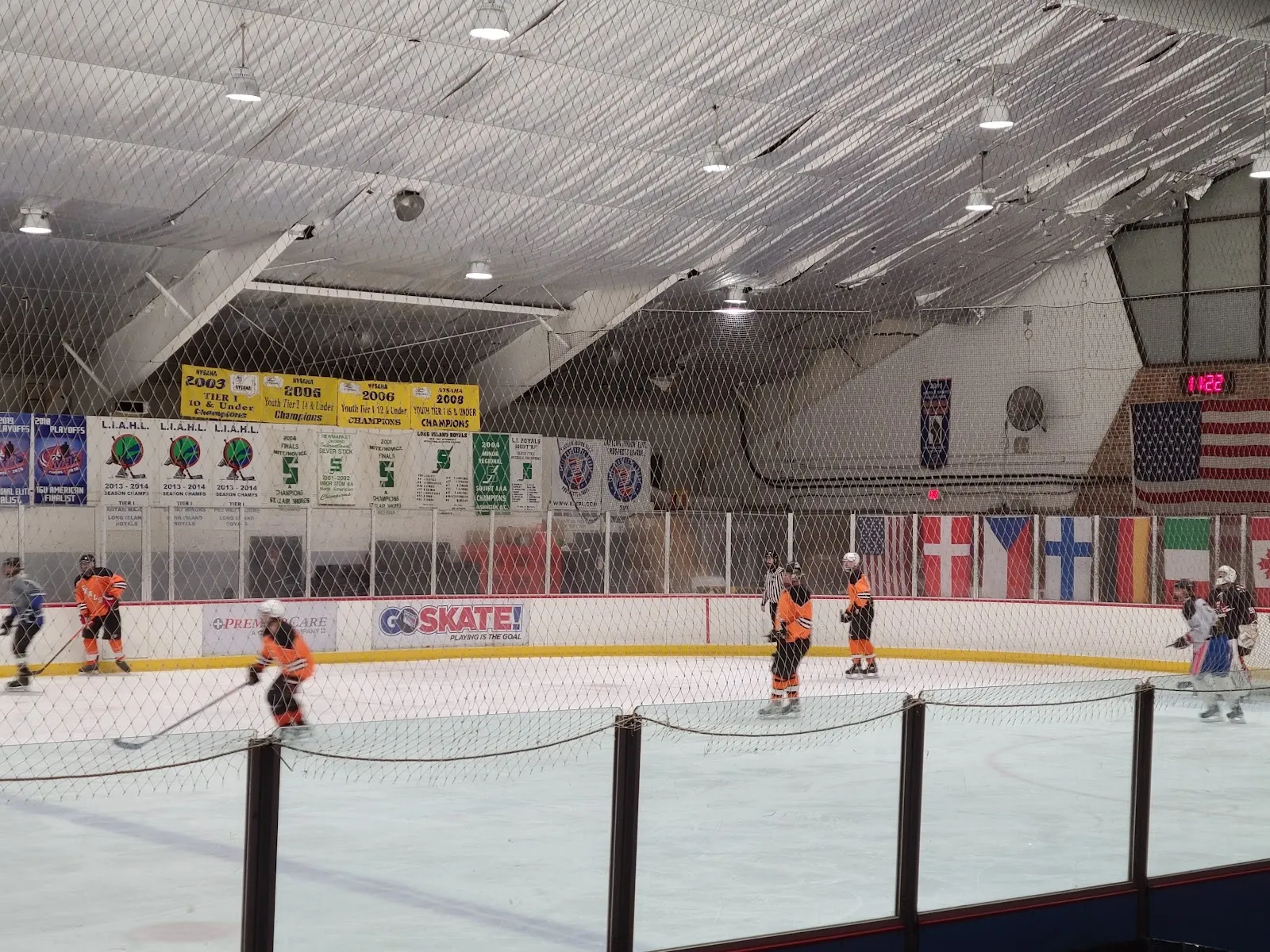 Un partido en curso en una pista bajo un techo de aislante blanco, con una bandera de Estados Unidos y banderas de varios países sobre la pared del fondo y una línea de vallas con cuerda al frente. — Superior Ice Rink