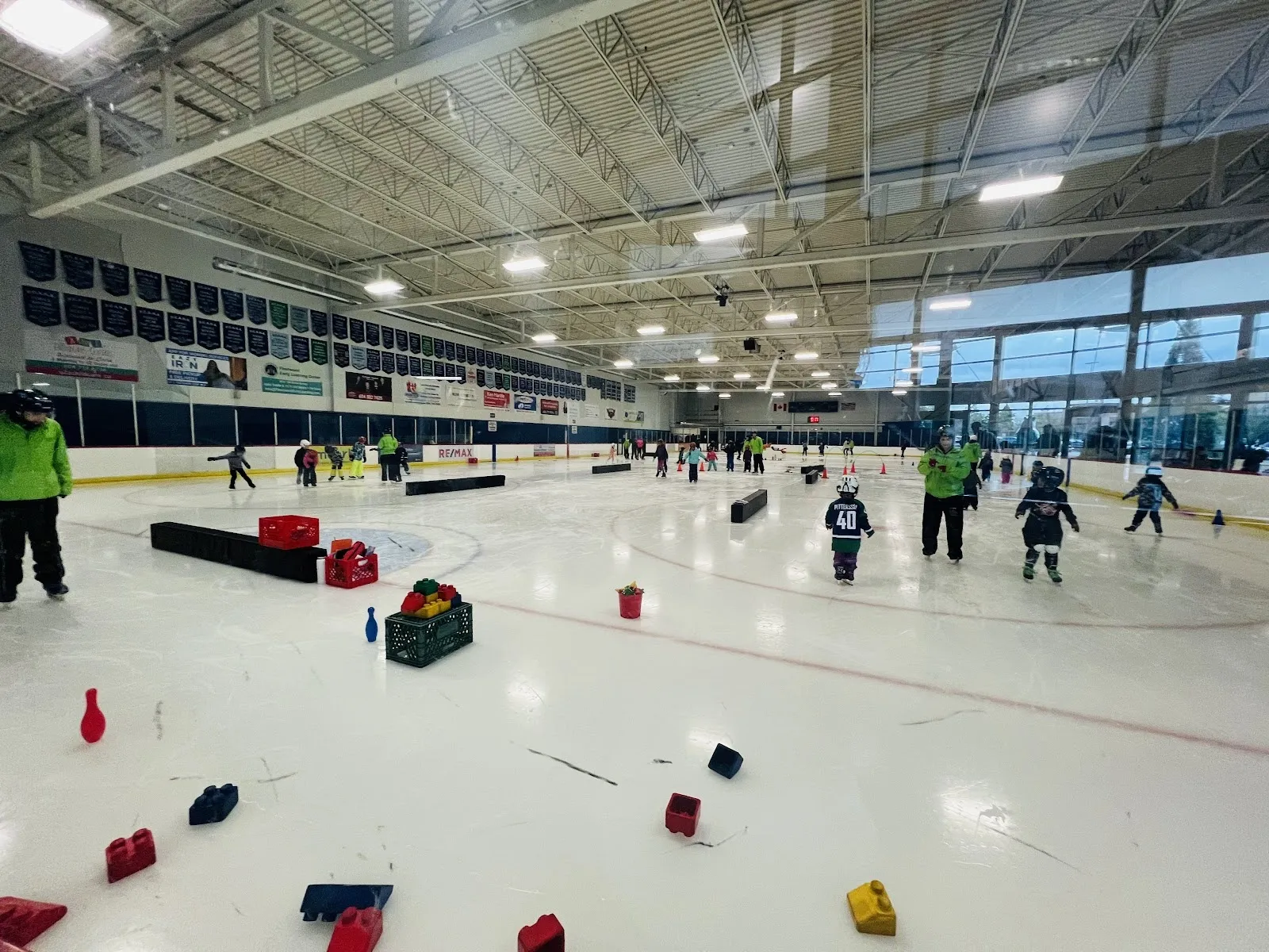 A learn-to-skate session on a wide sheet, skaters and instructors in green jackets spread among cones under a steel-trussed roof with a band of windows. — Surrey Sport & Leisure Complex Arena