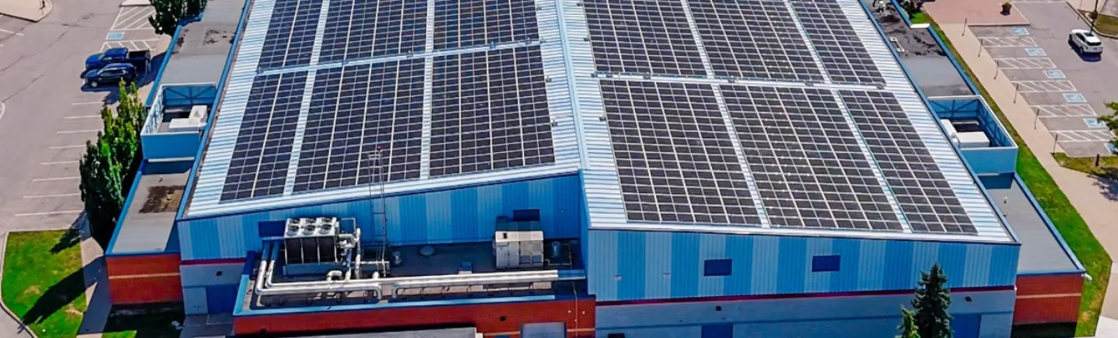Aerial view of the Susan Fennell Sportsplex building exterior, showing the blue-and-red metal-clad roofline covered in solar panels, rooftop mechanical units, and the surrounding parking lot. — Susan Fennell Sportsplex (South Fletcher's)