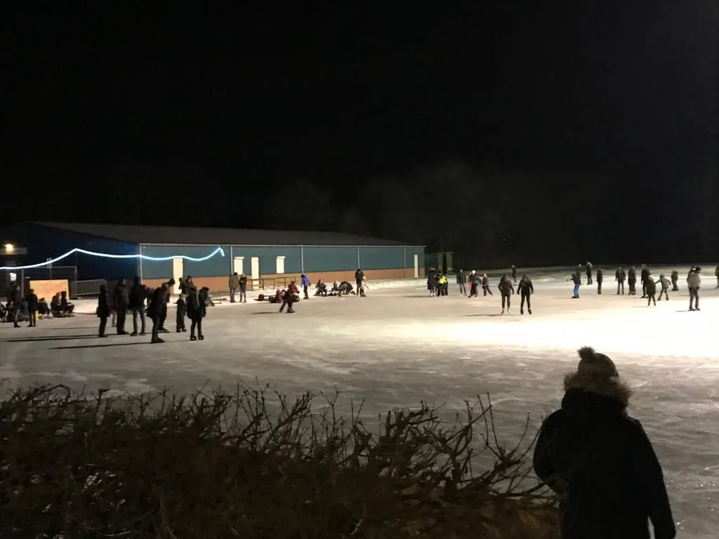 Skaters spread across the floodlit natural-ice rink at Bovenkarspel on an evening in February 2021, with the long clubhouse building and its string of lights at the left and two onlookers in the foreground. — Landijsbaan S.V. IJsvermaak (Bovenkarspel)