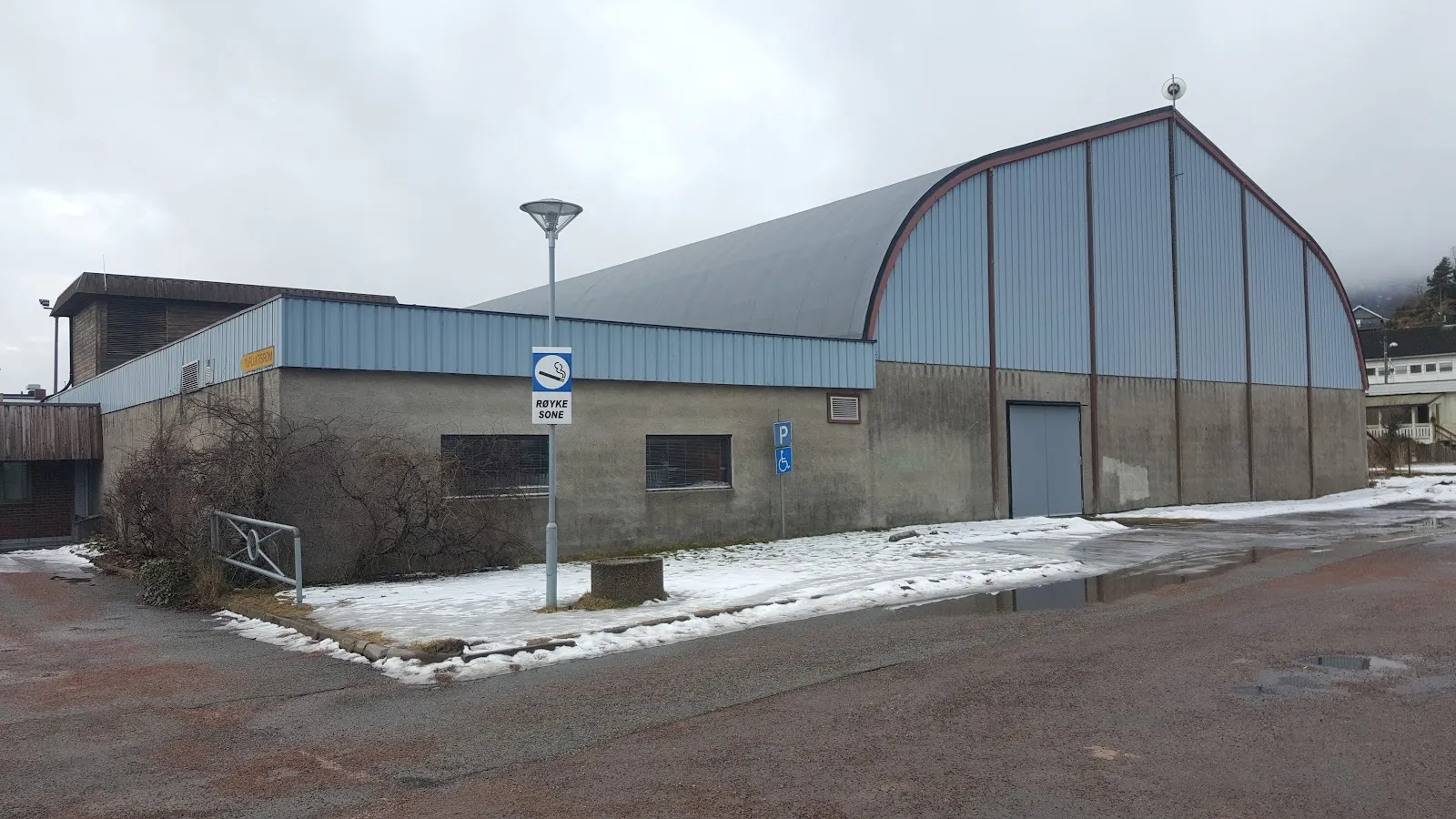The arched Strømmhallen sports hall in Svelvik with a snowy yard and a lamp post in front. — Svelvik kunstis (Strømmbanen)