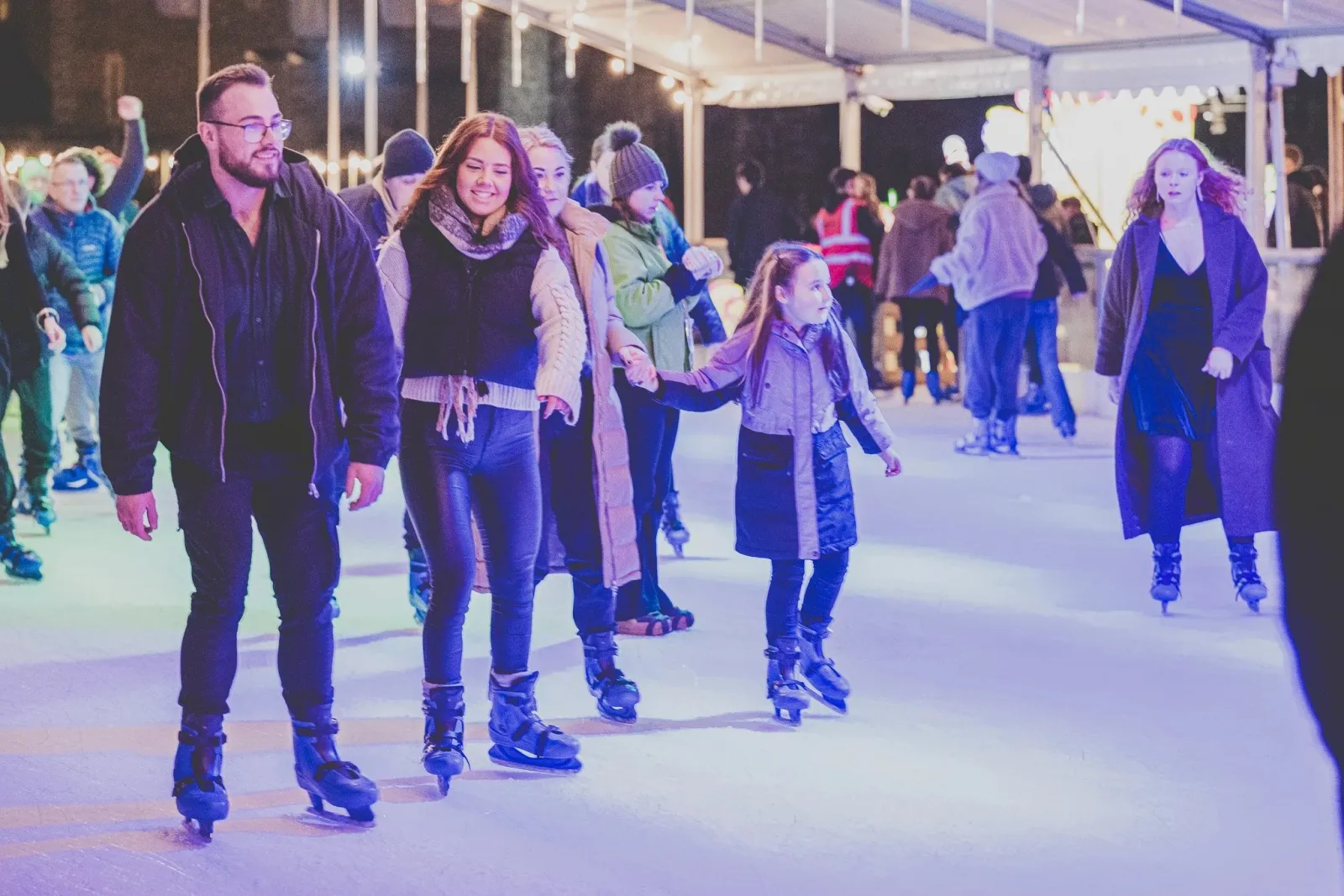 A line of skaters holding hands on the pale-lit ice at Swansea Waterfront Winter Wonderland, other skaters and the marquee roof behind. — Swansea Winter Wonderland Ice Rink