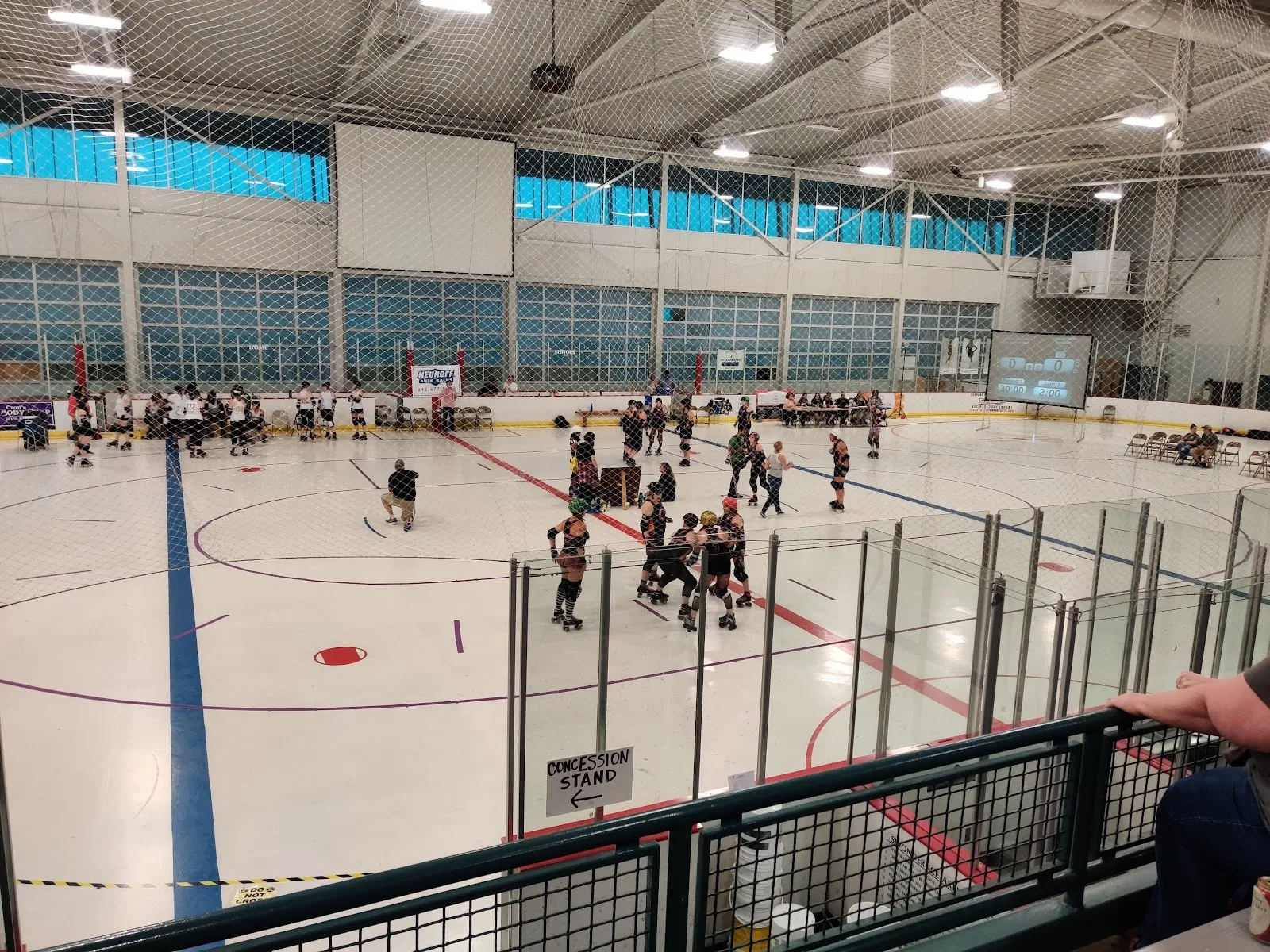 A team practice across a wide sheet seen from behind the glass, players clustered in the middle of the ice under a pale steel roof with a band of windows on the far wall. — Swonder Ice Arena