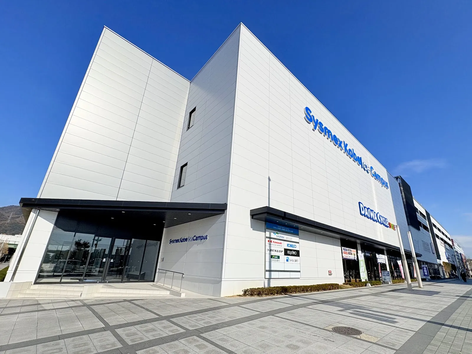 The white front of Sysmex Kobe Ice Campus under a clear blue sky, with the blue Sysmex Kobe Ice Campus lettering on the wall and the glass entrance at street level. — Sysmex Kobe Ice Campus