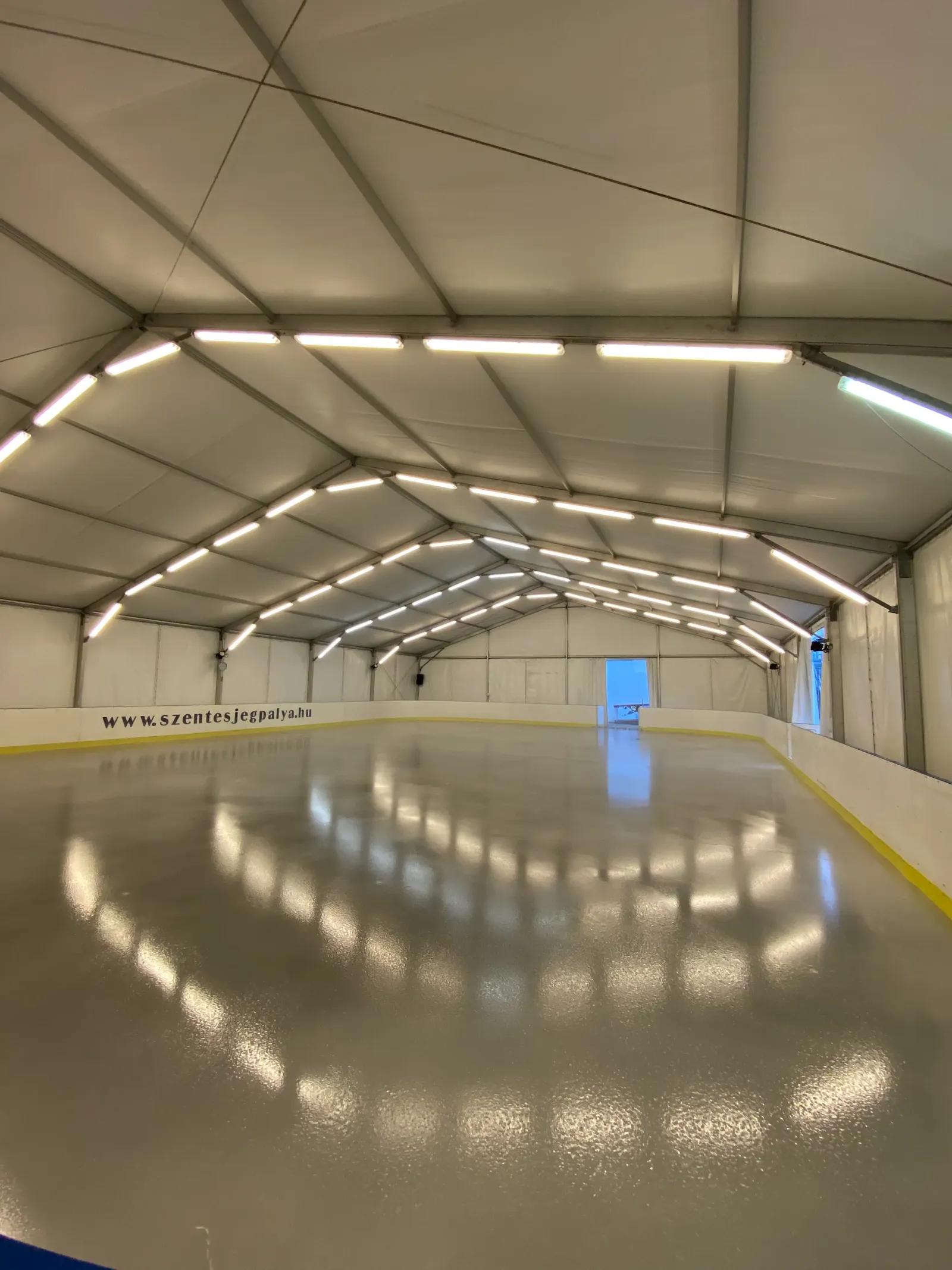 The empty ice under the tent roof of the Szentes Frigo ice rink, with the rink's web address along the boards — Szentes Frigo Jégpálya