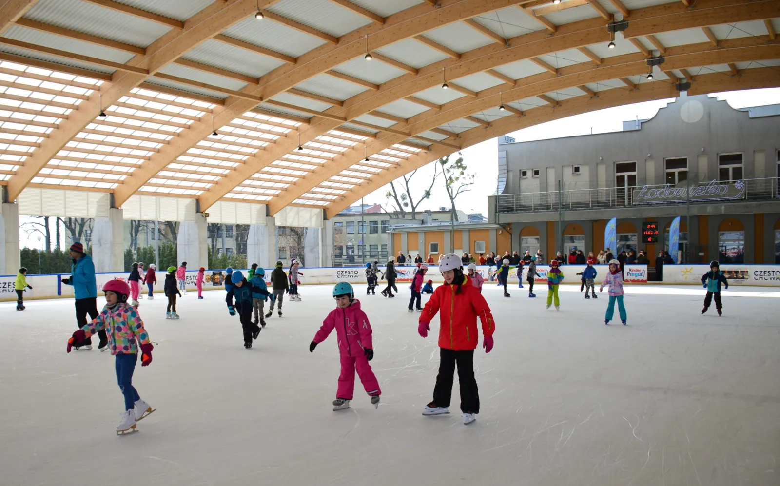 Children in helmets skating across the boarded ice under the glazed timber roof of the Częstochowa rink, with sponsor boards, a stand and the town's buildings seen through the open side. — Sztuczne Lodowisko MOSiR Częstochowa
