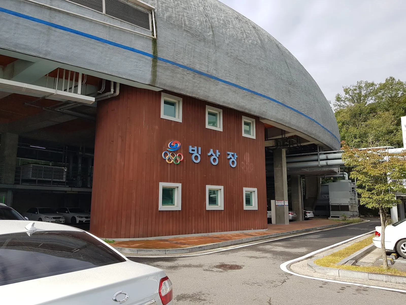 The ice rink building at the Korea National Training Center in Taereung: a curved concrete roof over a red timber wall carrying the Korean Olympic Committee emblem and the sign 빙상장 (ice rink), with the entrance road in front. — Taereung International Skating Rink