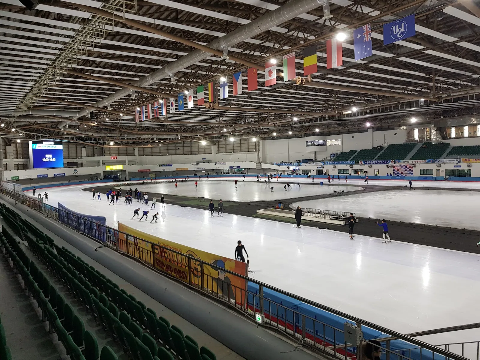 The speed-skating hall of the Korea National Training Center at Taereung: the long-track oval and the infield rink under the timber roof, with national flags along the rafters and a few skaters training in the distance. — Taereung International Skating Rink